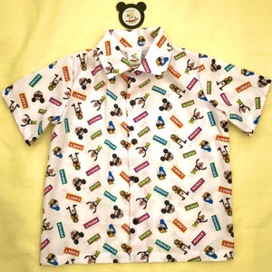 Może przedstawiać: Biała koszula z krótkim rękawem zapinana na guziki z kolorowym nadrukiem na całej powierzchni przedstawiającym Myszki Miki, Donalda Ducka, Goofy'ego i Pluto. Nadruk przedstawia postacie w okularach przeciwsłonecznych i kapeluszach z napisanymi pod nimi imionami.