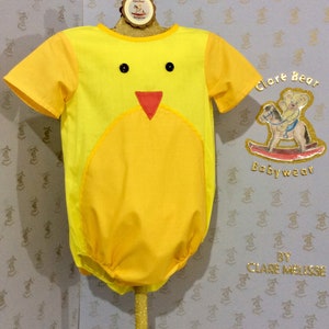May include: A yellow baby onesie with a chick applique. The onesie has a round yellow belly and a red beak. The onesie is on a stand with a sign that says "Clare Bear Babywear by Clare Melisse".