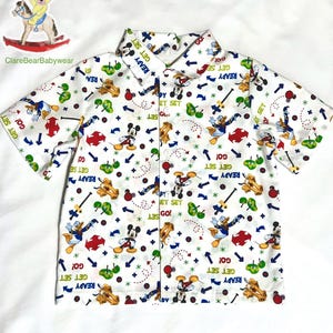 Könnte beinhalten: Weißes, kurzärmeliges Hemd mit Knöpfen und einem bunten Druck von Mickey Mouse, Donald Duck und anderen Disney-Figuren. Der Druck zeigt den Text "Get Set Go!" wiederholt.