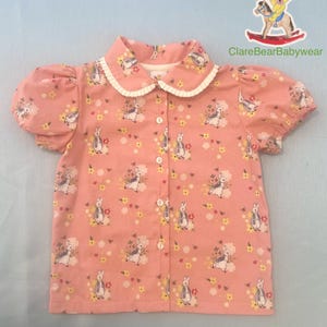 Puede incluir: Una camisa rosa de manga corta con botones, con estampado de Peter Rabbit y un cuello blanco Peter Pan. La camisa tiene cierre de botones y un ribete con volantes en el cuello.
