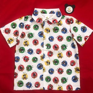 Könnte beinhalten: Weißes, kurzärmeliges Hemd mit Knöpfen und einem bunten Mickey-Mouse-Druck. Das Hemd hat einen Kragen und einen Knopfverschluss.
