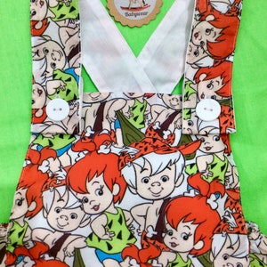 Puede incluir: Pantalón vaquero blanco para bebé con un diseño de tirantes cruzados. La tela presenta un estampado colorido de personajes de dibujos animados de los Picapiedra.