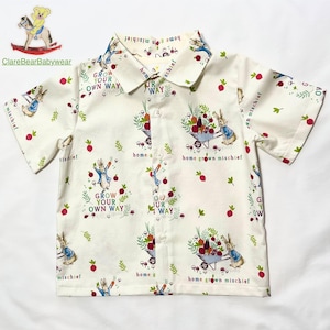Puede incluir: Camisa blanca de manga corta con botones con estampado de Peter Rabbit. El estampado presenta a Peter Rabbit con una carretilla llena de verduras, manzanas rojas y el texto "Grow Your Own Way" y "Home Grown Mischief".