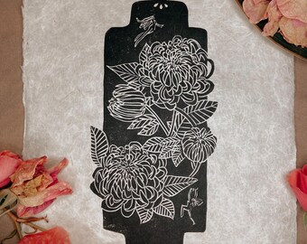 May include: Black and white floral linocut print on textured paper. The print features a stylized floral design within a rectangular shape with a decorative top and bottom. Dried roses and petals surround the print.