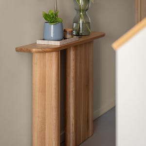 Può includere: Un tavolo console in legno con due gambe, con un piano curvo e una finitura in legno chiaro. Il tavolo è decorato con un vaso blu con una pianta verde, un vaso di vetro con fiori viola e un piccolo oggetto decorativo.