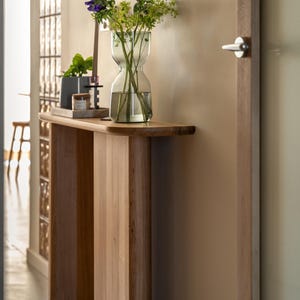 Op de afbeelding: Een lichtgekleurde houten console met een gebogen bovenkant. Een heldere glazen vaas met bloemen staat op de tafel, samen met een kleine potplant en een kaars. De muur achter de tafel is neutraal van kleur.