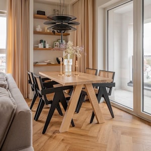 Op de afbeelding: Een houten eettafel met een lichte houtkleur en zwarte stoelen. De tafel heeft een uniek A-frame onderstel en is gedekt met een vaas met bloemen en kaarsen. De tafel staat in een moderne eetkamer met grote ramen en beige gordijnen.