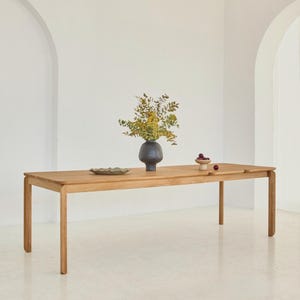 Peut inclure: Une table à manger en bois clair, avec un plateau rectangulaire et quatre pieds. Un vase bleu foncé avec de la verdure est posé sur la table, ainsi qu'une petite assiette et un bol de fruits. La table est dans une pièce blanche et lumineuse.