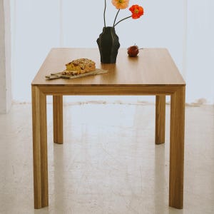 Puede incluir: Mesa cuadrada de madera clara. Un jarrón negro con flores naranjas y rojas, un pan sobre una tabla de madera y una manzana roja están sobre la mesa. La mesa tiene cuatro patas cuadradas. La mesa mide unos 76 cm de alto.