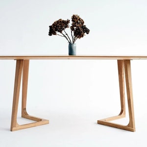 Puede incluir: Una mesa de comedor de madera clara con una tapa rectangular y patas únicas curvadas. La mesa está colocada sobre un fondo blanco con un jarrón de flores secas en la parte superior.