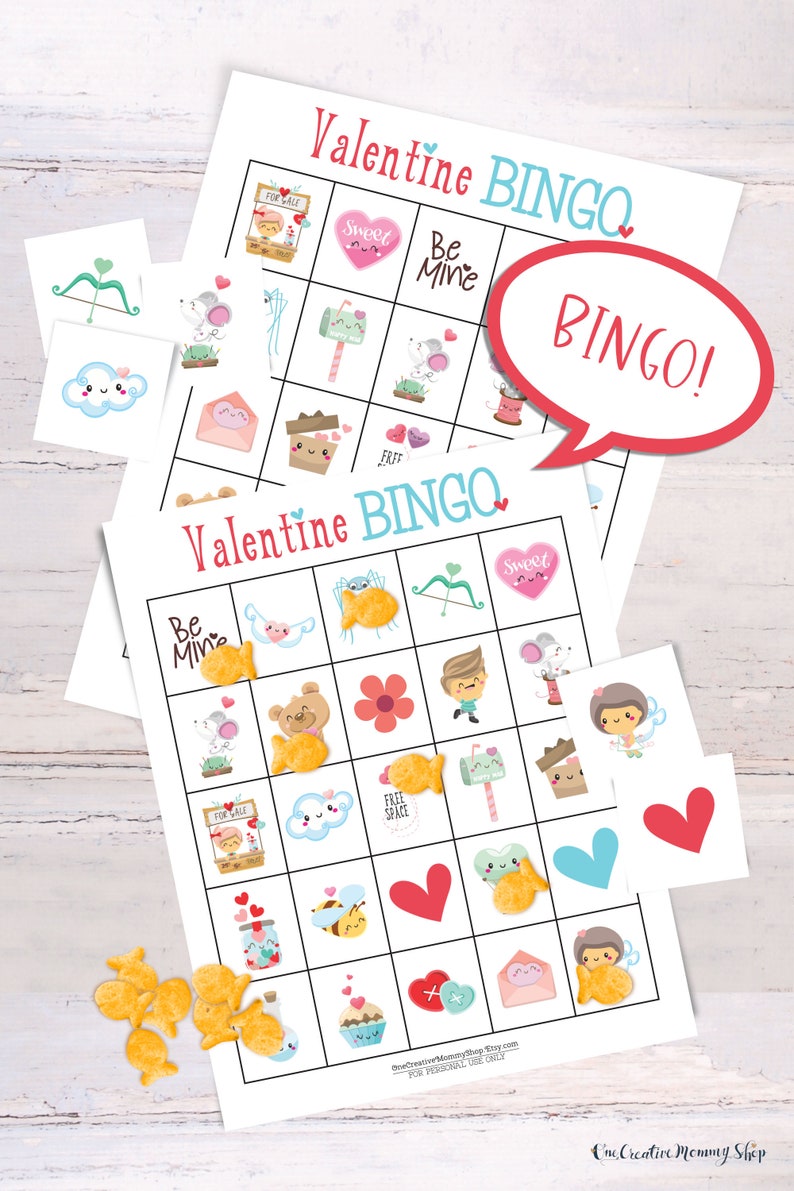 2 Valentines Day bingo cards are laid on top of each other  on a white table. There is a small pile of goldfish crackers. Six calling cards are scattered across the game, and several spots are marked with crackers. A word bubble says Bingo!