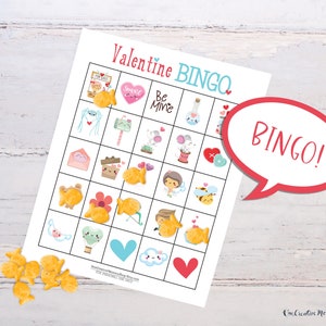 A cute Valentines Day bingo card is laid out on a white wooden table. There is a pile of goldfish crackers on the table, and several cover spaces on the bingo card. Five spots in a row are covered, and there is a word bubble that reads, Bingo!