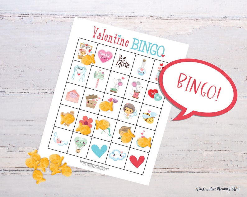 A cute Valentines Day bingo card is laid out on a white wooden table. There is a pile of goldfish crackers on the table, and several cover spaces on the bingo card. Five spots in a row are covered, and there is a word bubble that reads, Bingo!