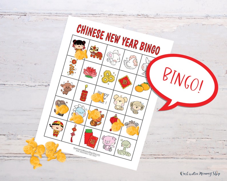 Chinese New Year bingo card lying on a white wooden table. Goldfish crackers are lying to the side of the board with several marking spaces on the bingo card. Five places are marked in a row, so there is a word bubble that reads, Bingo!