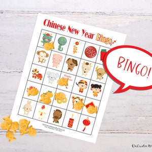 Lunar New Year bingo card lying on a white wooden table. Goldfish crackers are lying to the side of the board with several marking spaces on the bingo card. Five places are marked in a row, so there is a word bubble that reads, Bingo!