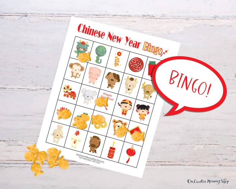 Lunar New Year bingo card lying on a white wooden table. Goldfish crackers are lying to the side of the board with several marking spaces on the bingo card. Five places are marked in a row, so there is a word bubble that reads, Bingo!