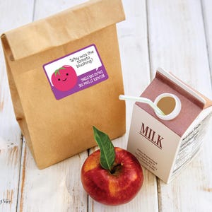 A brown paper lunch sack sits next to a red apple and a carton of milk with a straw sticking out. The lunchbox joke sticker on the front of the bag shows a cartoon tomato and says: Why was the tomato blushing? Because it saw the salad dressing.