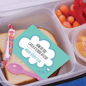 A closeup photo of an open insulated lunch bag with a bento container on top. It contains a sandwich, vegetables and fruit. On top of the sandwich is a lunchbox joke and a pack of smarties candy. How do you catch a school of fish? (With a book worm!)