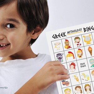 A young boy wearing a white T-shirt and missing 2 front teeth smiles while proudly holding up a Greek mythology bingo card.  Images include Zeus, Aphrodite, Ares, Poseidon, Hades, Ares, Hephaestus, Athena, Cerberus, Demeter, Hermes, Hera, Dionysus, Artemis, Pegasus, Apollo, and more.