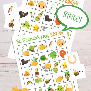 2 St. Patricks Day bingo cards are laid on top of each other on a white table. There is a small pile of goldfish crackers. Six calling cards are scattered across the game, and several spots are marked with crackers. A word bubble says Bingo!