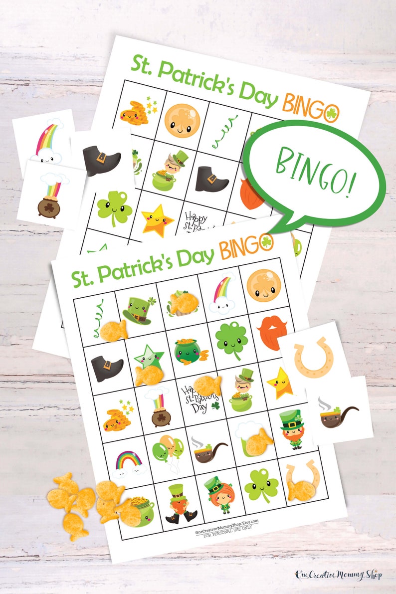 2 St. Patricks Day bingo cards are laid on top of each other on a white table. There is a small pile of goldfish crackers. Six calling cards are scattered across the game, and several spots are marked with crackers. A word bubble says Bingo!