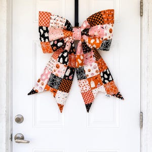 May include: A large decorative bow made of patchwork fabric featuring a Halloween theme. The fabric patterns include ghosts, pumpkins, spider webs, and checkered squares in orange, black, pink, and white. The bow is attached to a black door hanger.