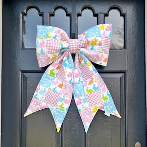 May include: A large, decorative bow made of patchwork fabric, featuring pastel pink, blue, and white squares with Easter bunny and egg motifs. The bow is attached to a black door, creating a festive spring or Easter decoration.
