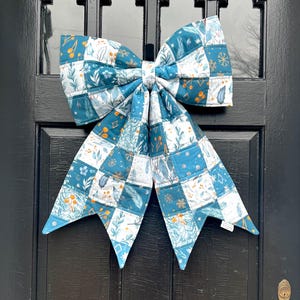 May include: A large, quilted fabric bow in shades of teal, white, and blue, adorned with floral and leaf patterns. The bow is attached to a black door, creating a festive and decorative accent.