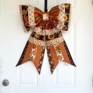 May include: A large decorative bow made from patchwork fabric featuring autumn-themed patterns including sunflowers, orange and brown floral prints, and plaid designs. The bow has two wide loops and two long, pointed tails hanging downwards.