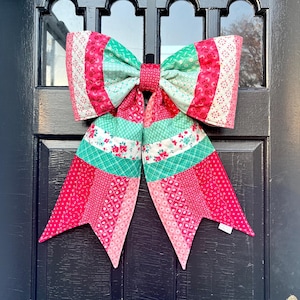 May include: A large, colorful fabric bow, likely for door decor. The bow features a patchwork design with shades of pink, red, and green. The bow is attached to a black door, creating a striking contrast. The bow's design includes floral and geometric patterns.