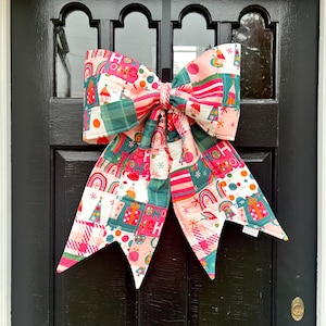 May include: A large, colorful fabric bow with a patchwork design, featuring Christmas-themed patterns like trees, rainbows, and gifts. The bow is predominantly pink, green, and white, and is hanging on a black door.