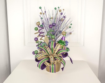 May include: A festive Mardi Gras centerpiece in a gold pot. It features purple, green, and gold ribbons, decorative sprigs, and ornaments. A party hat and spiral decorations add to the celebratory theme.