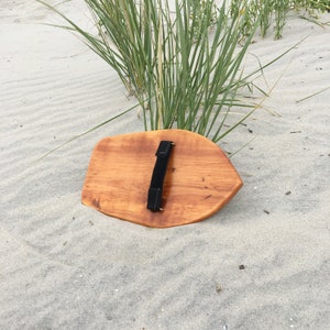 May include: A wooden shield with a black leather strap handle. The shield is lying on a sandy beach.
