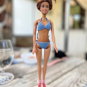 以下が含まれることがあります： 高い位置でお団子に結った茶色の髪のドールが、水色のクロシェ編みのツーピースビキニを着用しています。ドールは日焼けした肌にピンクの唇をしており、透明な円形のプラスチック製スタンドの上に立ち、足元には鮮やかなピンクの靴を履いています。