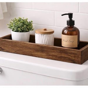 Può includere: Un vassoio di legno tinto scuro contiene una piccola pianta in vaso, un barattolo bianco con coperchio in legno e una bottiglia di vetro marrone di sapone per le mani. La bottiglia di sapone per le mani ha una pompa nera e un'etichetta con la scritta "Hand Soap".