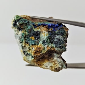 May include: A rough, irregularly shaped rock with a mix of green, blue, and brown colors. The rock has a variety of textures and appears to be a mineral specimen.