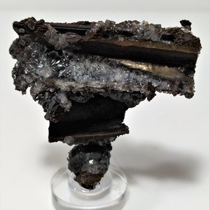 May include: A dark brown and silver mineral specimen with a layered structure. The mineral is mounted on a clear acrylic base.