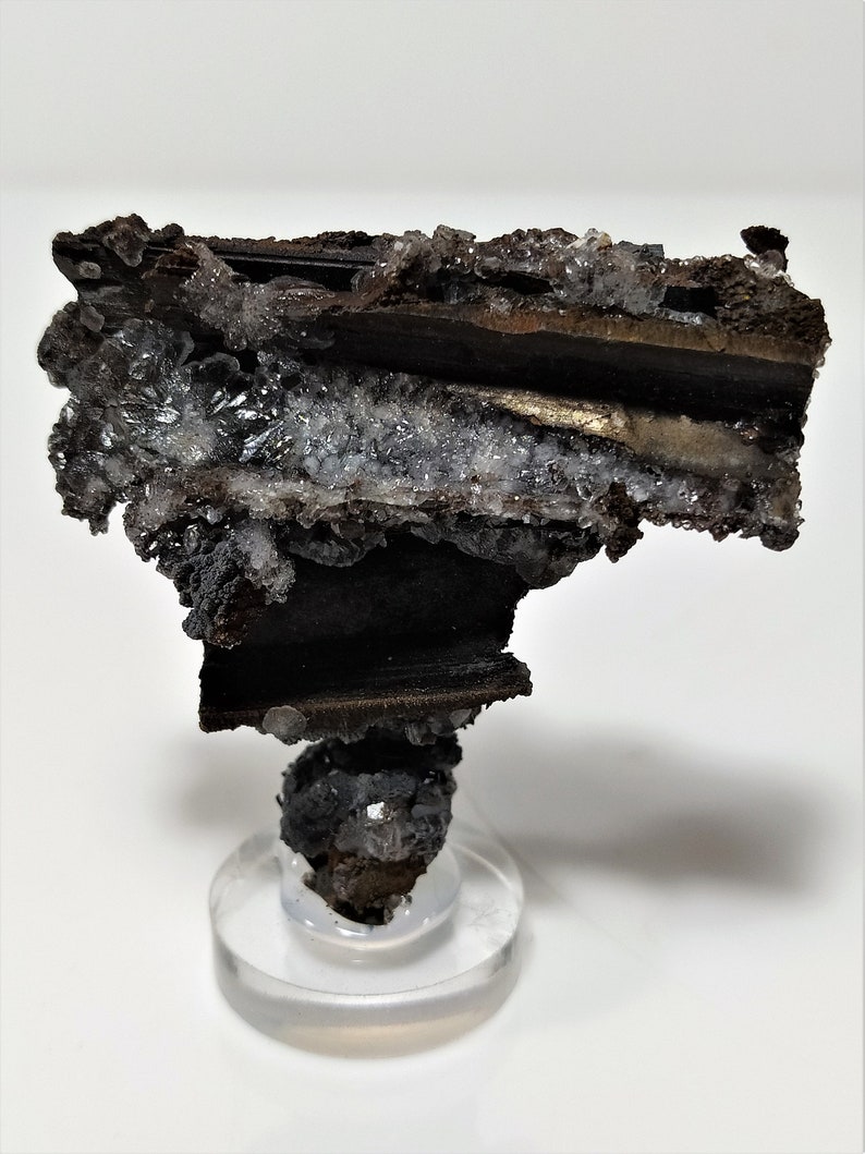 May include: A dark brown and silver mineral specimen with a layered structure. The mineral is mounted on a clear acrylic base.