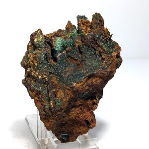 May include: A natural mineral specimen, displaying a rugged texture and a blend of brown and green colours. The rock sits on a clear acrylic stand. The surface of the mineral has a crystalline appearance.