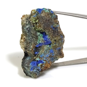 Può includere: Un masso ruvido di colore marrone scuro con depositi minerali blu e verdi. Il masso è tenuto da una pinzetta.