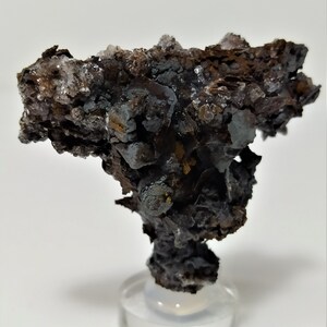 May include: A dark gray and brown mineral specimen with a rough, textured surface. The specimen is on a clear display stand.