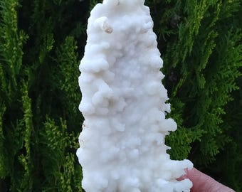 Op de afbeelding: Een witte, natuurlijk gevormde kristalkluster met een ruw, getextureerd oppervlak. De kristal is gevormd als een hoge, dunne driehoek.