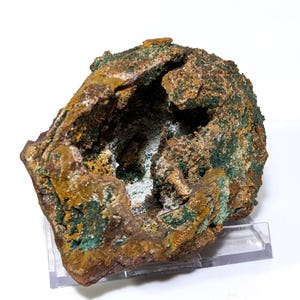 May include: A large, irregularly shaped rock with a hollow center. The rock is a mix of brown, orange, and green colors. It is displayed on a clear acrylic stand.