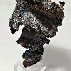 May include: A dark brown, metallic, rock-like formation with a rough, textured surface. The formation is covered in small, white, crystalline formations. The rock is displayed on a clear, round stand.