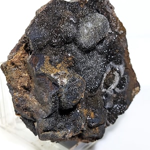 May include: A close-up of a dark gray and brown rock with a cluster of small, shiny, silver-colored crystals. The rock is on a clear acrylic stand.