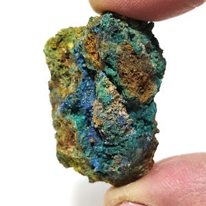 May include: A close-up of a mineral specimen held between fingers. The rock features a mix of blue, green, and brown hues. The texture appears rough and uneven, with visible crystalline structures. The mineral is likely a natural stone.