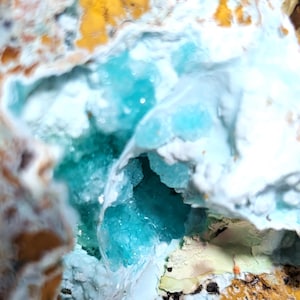 May include: Close-up of a geode with a vibrant turquoise crystal formation within a cavity. The outer rock has a mix of white, yellow, and brown hues, creating a striking contrast with the crystal's color. The geode's texture is rough and natural.