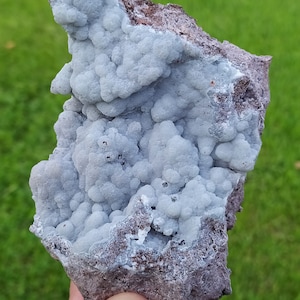 May include: A natural mineral specimen with a light blue, bubbly crystal formation on a dark brown and grey matrix. The crystal clusters have a rounded, textured appearance. The background is a blurred green lawn.