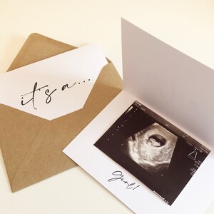 May include: A white card with the text "it's a..." on the front. The card is open to reveal a black and white ultrasound image with the text "girl!" below it.