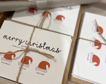 May include: A stack of Christmas cards featuring a red and white Santa hat design. The cards read "merry christmas" and have the names Thomas, Evita, and Oscar on them.